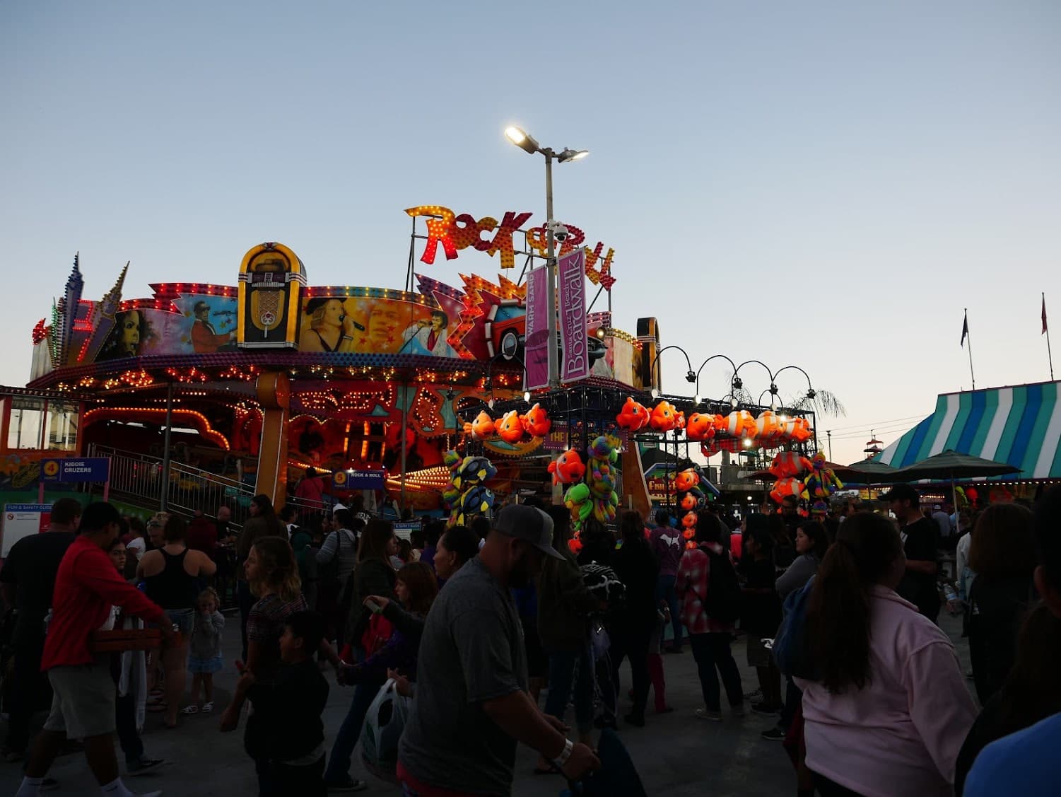 Crowded boardwalk midway beneath illuminated rides and rows of orange fish prizes.
