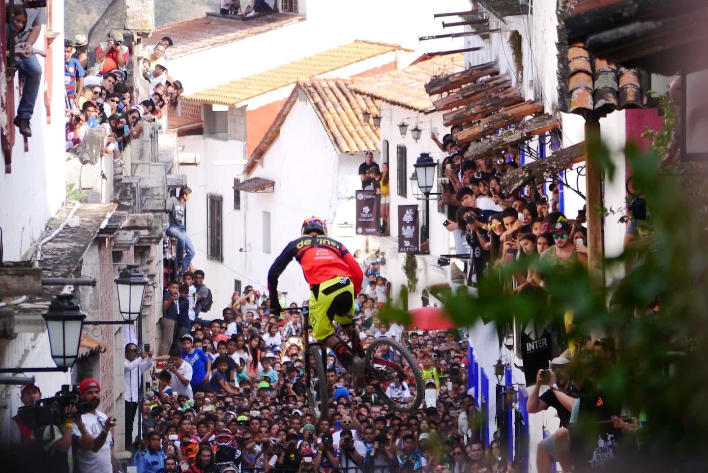 Cyclist jumping above a crowd on a narrow street lined with buildings.