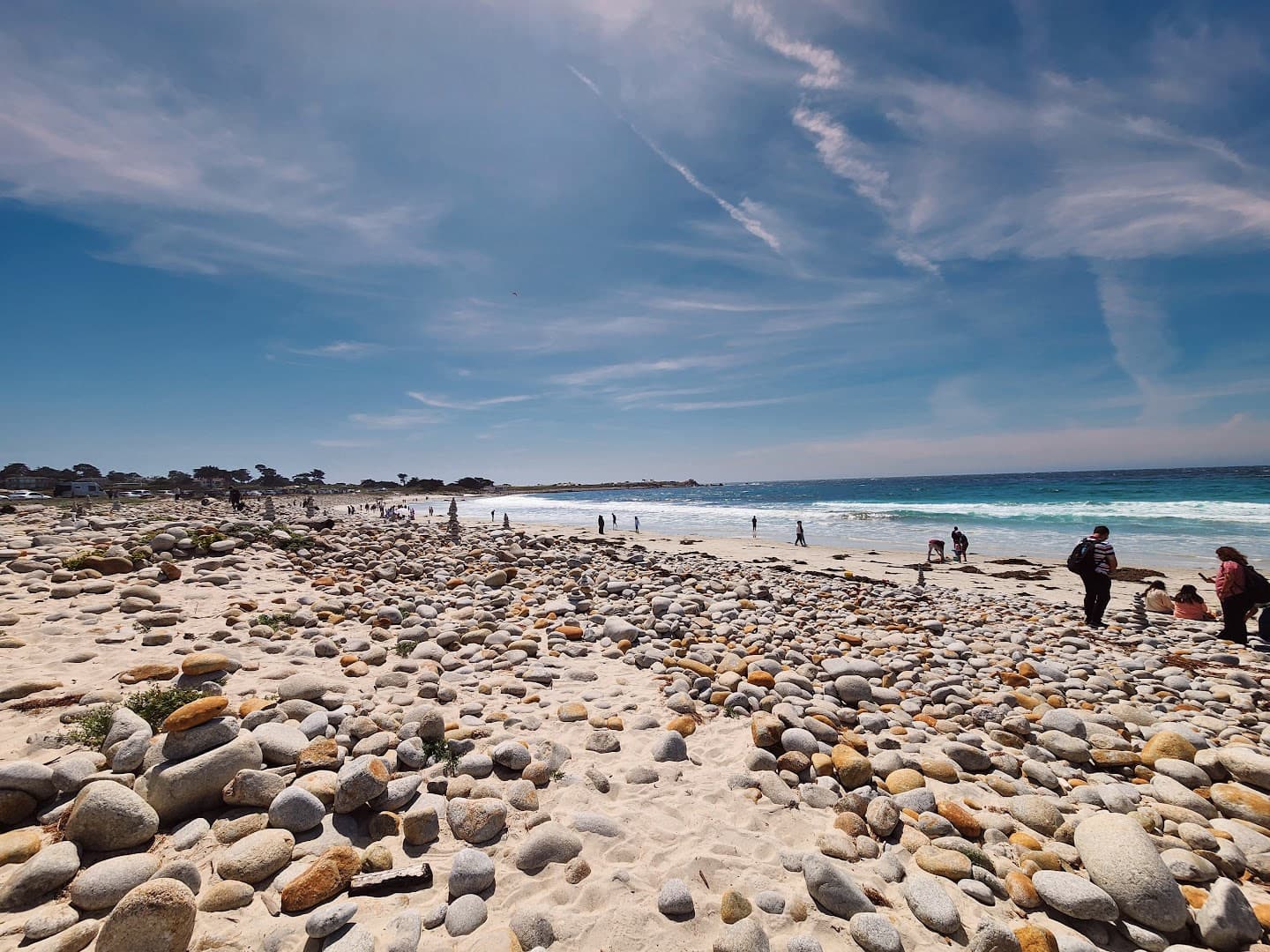 Rocky beach with people beside blue surf under a wide, streaked sky.