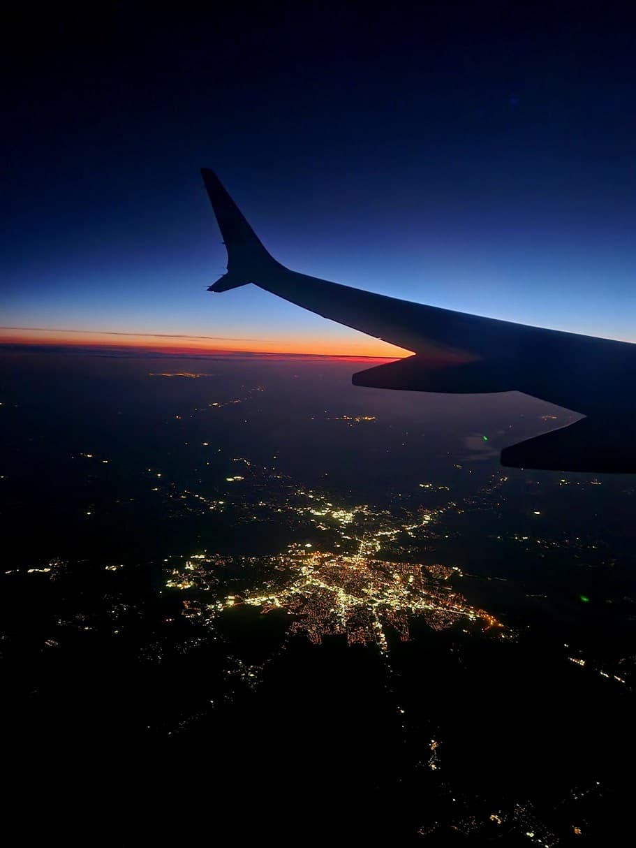 Airplane wing silhouetted above scattered city lights at sunset.