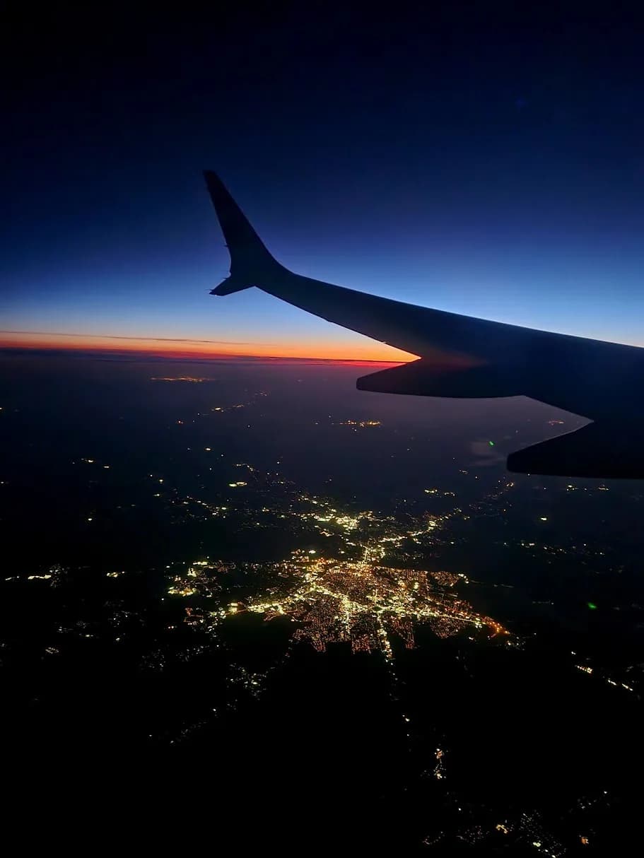 Airplane wing silhouetted above scattered city lights at sunset.